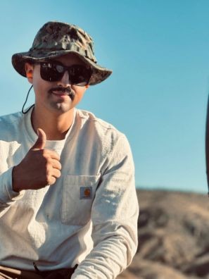 Person wearing sunglasses and a camo hat giving a thumbs-up outdoors with a clear sky.